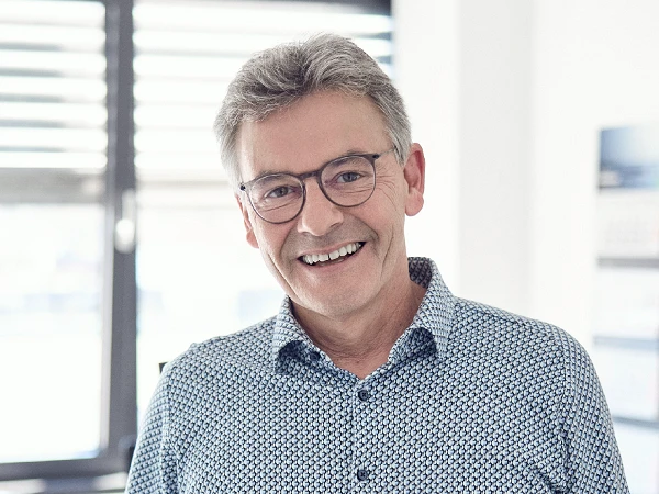 Ein lächelnder Mann mit grauen Haaren und Brille trägt ein hellblaues Hemd mit Muster und steht in einem hellen, modernen Büro.