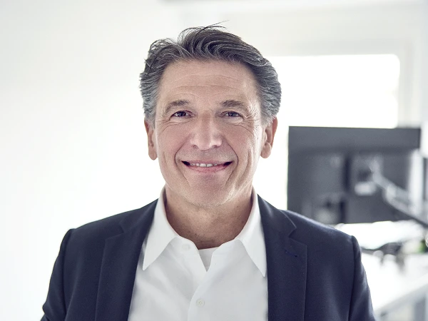 Ein Mann mittleren Alters mit kurzen, grauen Haaren lächelt freundlich in einem modernen Büro, das einen Schreibtisch und einen Computer im Hintergrund zeigt.
