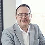 Ein lächelnder Mann mit Brille trägt einen hellgrauen Blazer und ein weißes Hemd, während er in einem gut beleuchteten Raum sitzt.