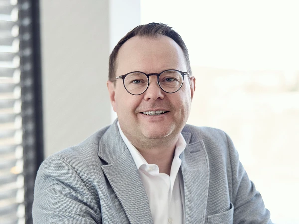 Ein lächelnder Mann mit Brille trägt einen hellgrauen Blazer und ein weißes Hemd, während er in einem gut beleuchteten Raum sitzt.
