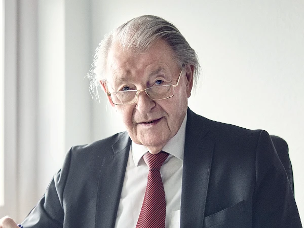 Ein älterer Mann mit schütterem grauem Haar und einer Brille, trägt einen schwarzen Anzug und eine rot gemusterte Krawatte. Er sitzt an einem Tisch und schaut direkt in die Kamera, während er einen Stift hält. Die Umgebung wirkt modern und hell.