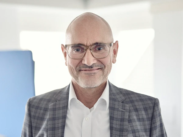 Ein lächelnder, älterer Mann mit Brille trägt einen karierten Anzug und ein weißes Hemd und steht in einem hellen Büro.