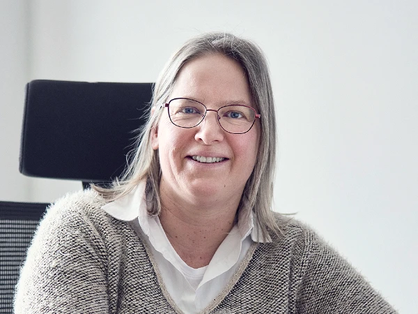 Eine lächelnde Frau mit langen, grauen Haaren und Brille sitzt an einem Schreibtisch, trägt einen hellen Pullover und schaut freundlich in die Kamera.