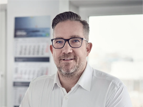 Ein lächelnder Mann mit Brille trägt ein weißes Hemd und steht in einem modernen Büro mit Fenstern, im Hintergrund sind Kalender und Dokumente sichtbar.