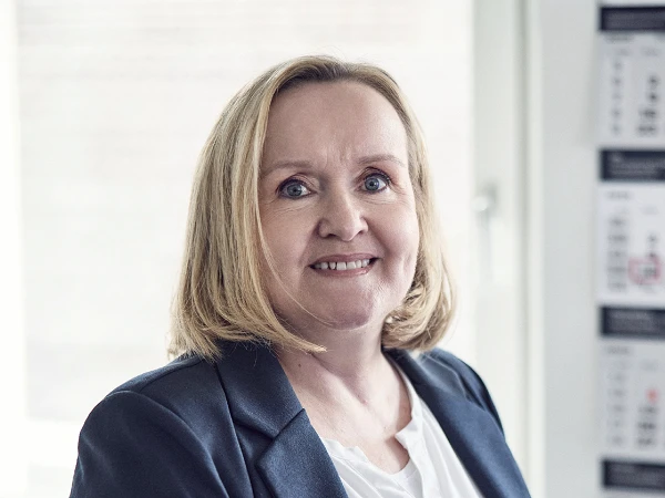 Eine lächelnde Frau mit blonden, schulterlangen Haaren trägt ein dunkelblaues Blazer und ein weißes Hemd. Sie steht vor einem hellen Hintergrund mit einigen unscharfen Drucken an der Wand.