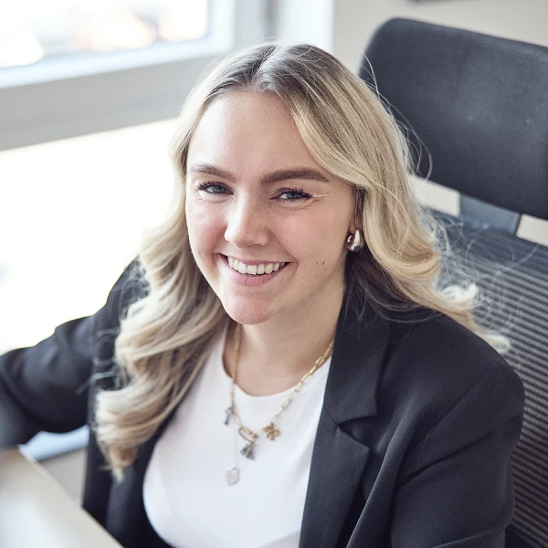Eine lächelnde Frau mit langen, blonden Haaren sitzt an einem Schreibtisch in einem modernen Büro und trägt ein schwarzes Blazer.