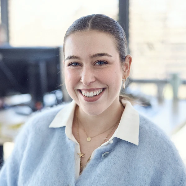 Eine lächelnde junge Frau mit braunen Haaren und blauer Strickjacke sitzt in einem modernen Büro, während sie in die Kamera schaut. Im Hintergrund sind mehrere Computerarbeitsplätze sichtbar.