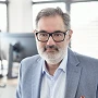 Ein Mann mit Brille und grauem Bart trägt einen hellen Anzug und ein gestreiftes Hemd. Er schaut direkt in die Kamera, während im Hintergrund ein moderner Arbeitsplatz mit Computern sichtbar ist.