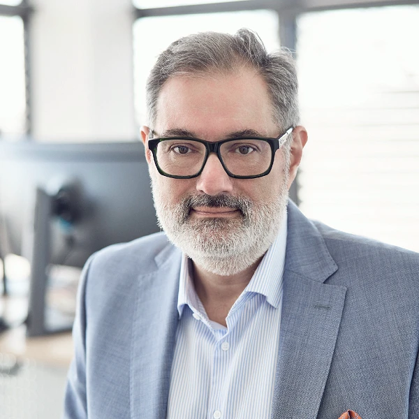 Ein Mann mit Brille und grauem Bart trägt einen hellen Anzug und ein gestreiftes Hemd. Er schaut direkt in die Kamera, während im Hintergrund ein moderner Arbeitsplatz mit Computern sichtbar ist.