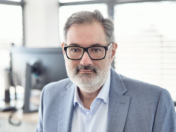 Ein Mann mit Brille und grauem Bart trägt einen hellen Anzug und ein gestreiftes Hemd. Er schaut direkt in die Kamera, während im Hintergrund ein moderner Arbeitsplatz mit Computern sichtbar ist.