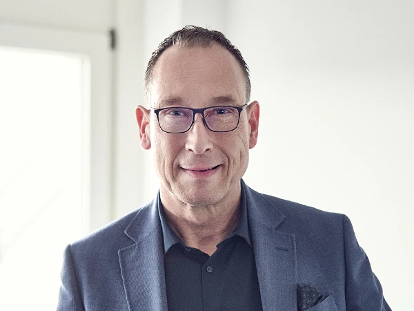 Ein Mann mit Brille und kürzeren, dunklen Haaren trägt einen blauen Blazer und steht vor einem Fenster, während er freundlich in die Kamera lächelt.