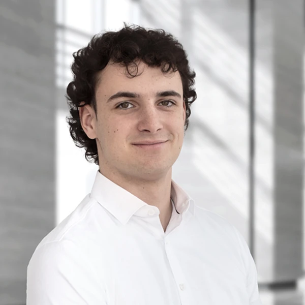Ein junger Mann mit lockigem Haar trägt ein weißes Hemd und steht in einem modernen, hellen Innenraum mit großen Fenstern.
