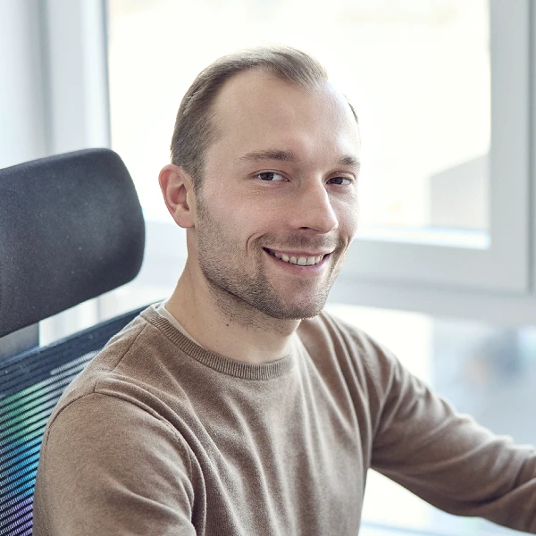 Ein Mann in einem hellbraunen Pullover lächelt freundlich in die Kamera, während er in einem modernen Bürostuhl sitzt, mit großen Fenstern und hellem Licht im Hintergrund.