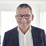 Ein lächelnder Mann mit kurzen, grauen Haaren und Brille trägt einen dunklen Blazer und ein weißes Hemd. Er steht in einem hellen Raum mit unscharfen Möbeln im Hintergrund.