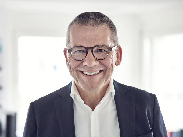 Ein lächelnder Mann mit kurzen, grauen Haaren und Brille trägt einen dunklen Blazer und ein weißes Hemd. Er steht in einem hellen Raum mit unscharfen Möbeln im Hintergrund.