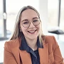 Eine lächelnde Frau mit langen, glatten Haaren sitzt in einem modernen Büro. Sie trägt eine orangefarbene Blazerjacke und eine blaue Bluse, während sie durch ihre Brille blickt. Im Hintergrund sind hell erleuchtete Fenster zu sehen.