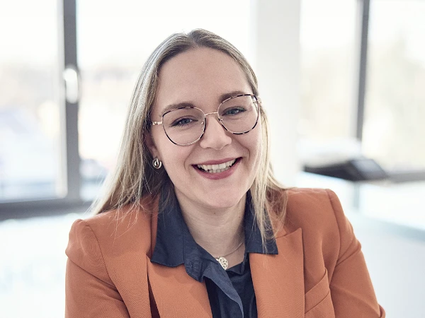 Eine lächelnde Frau mit langen, glatten Haaren sitzt in einem modernen Büro. Sie trägt eine orangefarbene Blazerjacke und eine blaue Bluse, während sie durch ihre Brille blickt. Im Hintergrund sind hell erleuchtete Fenster zu sehen.