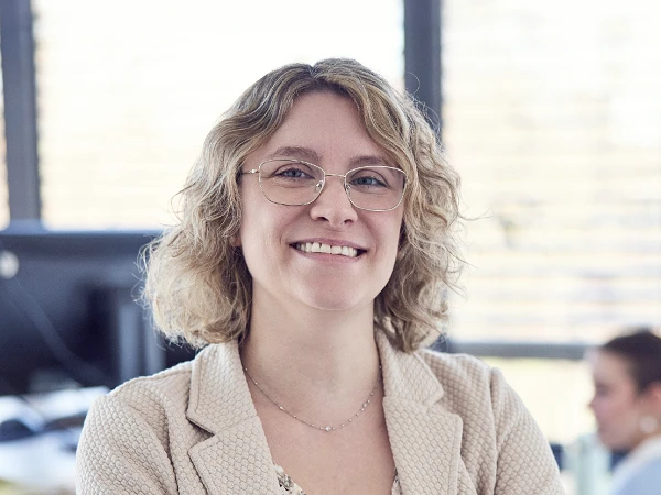Eine lächelnde Frau mit lockigem, blondem Haar und Brille steht im Vordergrund eines modernen Büros, während im Hintergrund weitere Mitarbeitende zu sehen sind.
