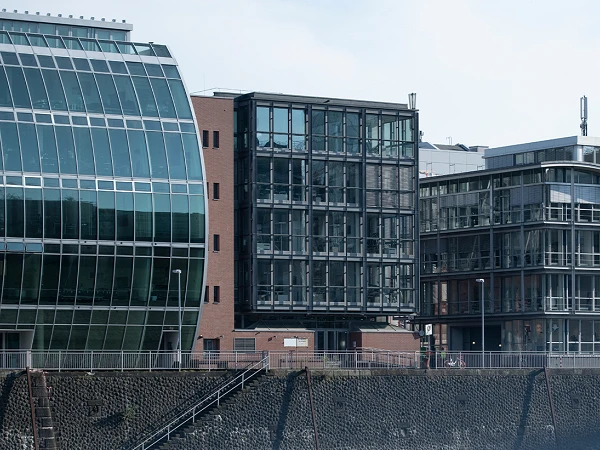 Moderne Gebäude mit unterschiedlichen architektonischen Stilen, darunter große Glasfronten und gewellte Fassaden, stehen entlang eines Ufers.