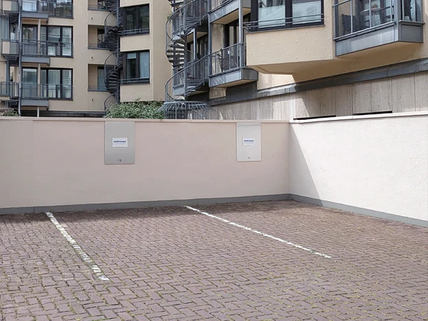 Leerer Parkplatz mit grauer Pflasterung, umgeben von hellen Gebäuden mit Balkonen und einer Wendeltreppe im Hintergrund. An der Wand sind zwei graue Kästen angebracht.
