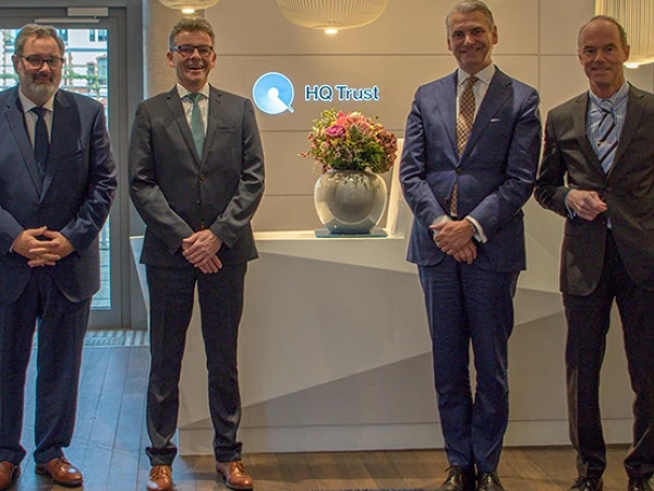 Vier Geschäftsleute in formeller Kleidung stehen nebeneinander vor einem Empfangstresen mit dem Logo von HQ Trust und einer Blumenvase.
