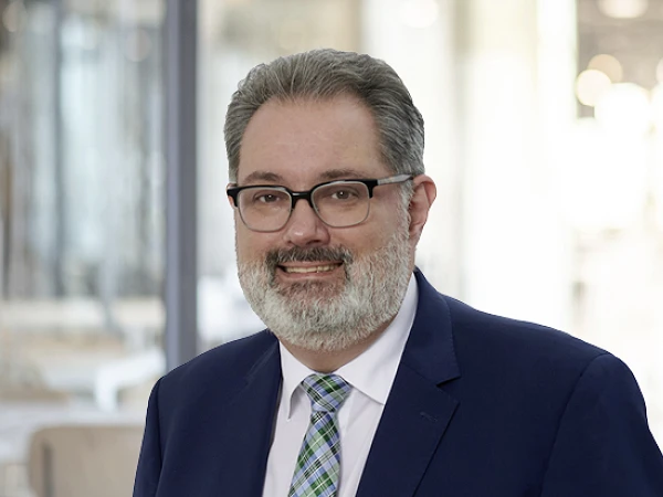 Ein Mann mit Brille und grauem Bart trägt einen dunklen Anzug und ein helles Hemd mit einer karierten Krawatte. Er lächelt freundlich vor einem unscharfen Hintergrund, der eine moderne Innenarchitektur zeigt.
