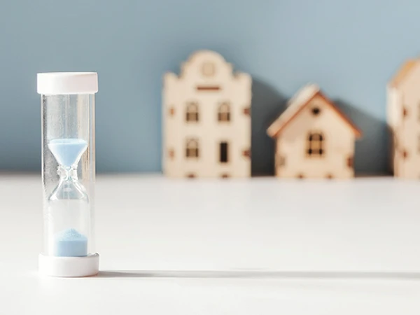 Eine Sanduhr mit blauem Sand steht auf einem Tisch, während im Hintergrund mehrere kleine, filigrane Holzmodelle von Häusern zu sehen sind.
