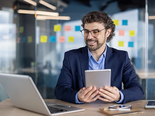 Ein lächelnder Mann mit Brille sitzt an einem Tisch und hält ein Tablet in der Hand, während er auf einen Laptop vor sich schaut. Im Hintergrund sind bunte Haftnotizen an einer Wand angebracht.