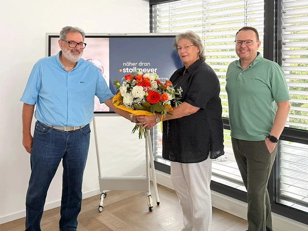 Eine Person überreicht einer anderen Person einen großen Blumenstrauß, während ein Dritter lächelnd daneben steht. Im Hintergrund ist ein Bildschirm mit dem Text „näher dran • stoll + never“ zu sehen.