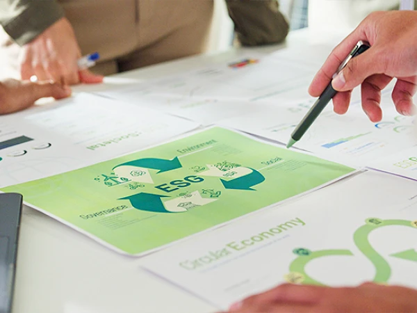 Auf einem Tisch liegen mehrere Dokumente, darunter eine Grafik mit dem Titel "ESG" und Symbolen, die soziale, ökonomische und ökologische Aspekte darstellen. Eine Hand mit einem Stift zeigt auf die Grafik, während andere Hände und Stifte im Hintergrund sichtbar sind.
