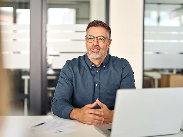 Ein Mann mit Brille und kurzem, grauem Bart sitzt an einem Schreibtisch in einem modernen Büro und spricht. Vor ihm steht ein Laptop, und er wirkt freundlich und professionell.