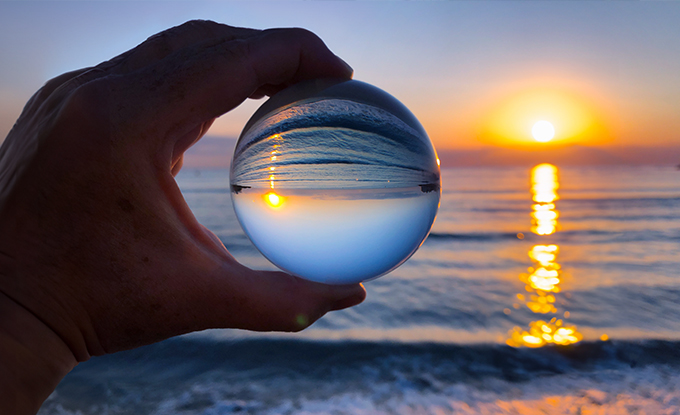 Eine Hand hält eine Glaskugel, in der der Sonnenuntergang über dem Meer reflektiert wird, während die Wellen sanft am Strand plätschern.