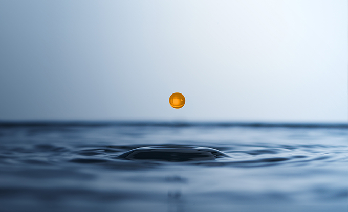 Eine orangefarbene Tropfen schwebt über der Wasseroberfläche und erzeugt Wellenripple, während der Hintergrund sanfte Blau- und Grautöne zeigt.