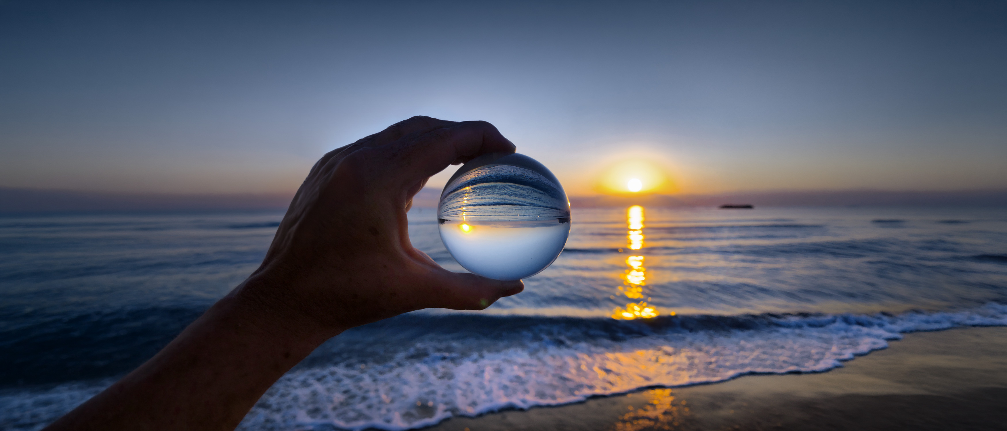 Eine Hand hält eine Glaskugel, die den Sonnenuntergang über dem Meer reflektiert, während sanfte Wellen an den Strand rollen.