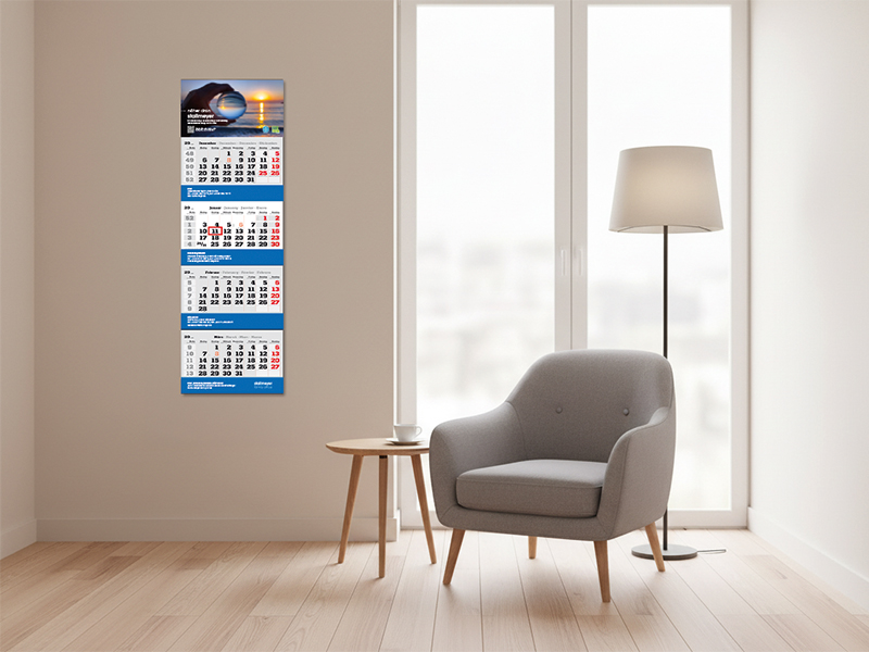 Ein Wandkalender mit mehreren Monaten ist an einer hellen Wand befestigt, während ein grauer Sessel und eine Stehlampe im Raum stehen. Ein Fenster lässt Tageslicht herein.
