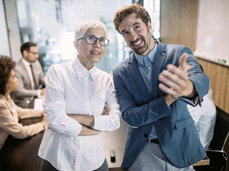 Ein lächelndes männliches und weibliches Teammitglied stehen in einem modernen Besprechungsraum, während sie mit der Hand auf einen gemeinsamen Gesprächspartner zeigen. Im Hintergrund sind weiteren Personen an einem Tisch zu sehen, die anscheinend an einem Meeting teilnehmen.