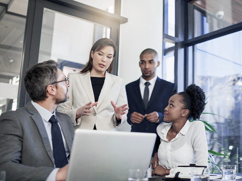 Eine Businessbesprechung in einem modernen Büro, bei der eine Frau im Anzug mit gestikulierenden Händen eine Präsentation hält, während zwei Personen am Tisch aufmerksam zuhören. Ein Mann steht im Hintergrund und beobachtet das Gespräch.