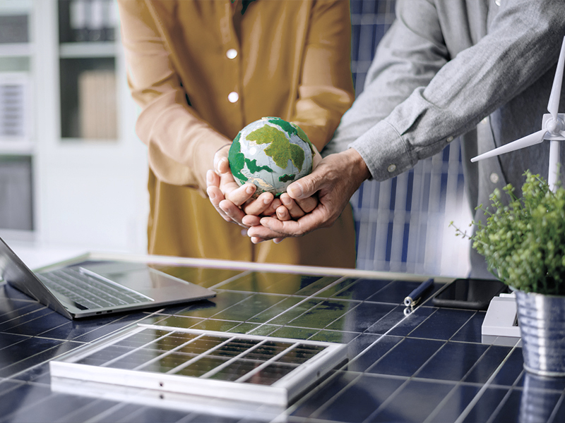Zwei Personen stehen an einem Tisch, auf dem Solarpanels und ein kleiner Topf mit einer Pflanze liegen, während sie gemeinsam eine kleine, grüne Erdkugel halten.