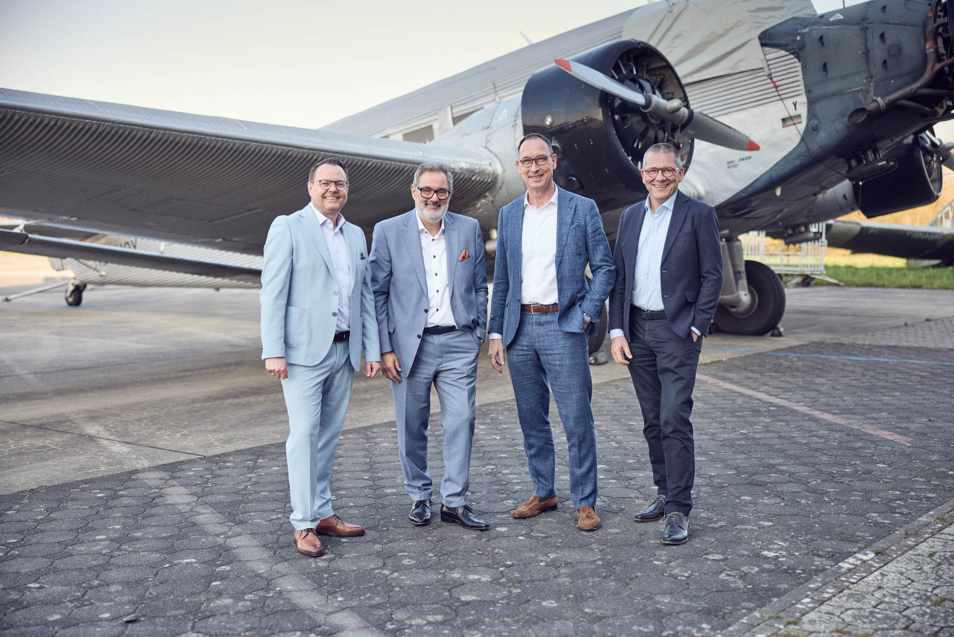 Vier Männer in eleganter Kleidung posieren lächelnd vor einem historischen Flugzeug auf einer gepflasterten Fläche. Das Flugzeug befindet sich im Hintergrund, und die Umgebung wirkt hell und freundlich.