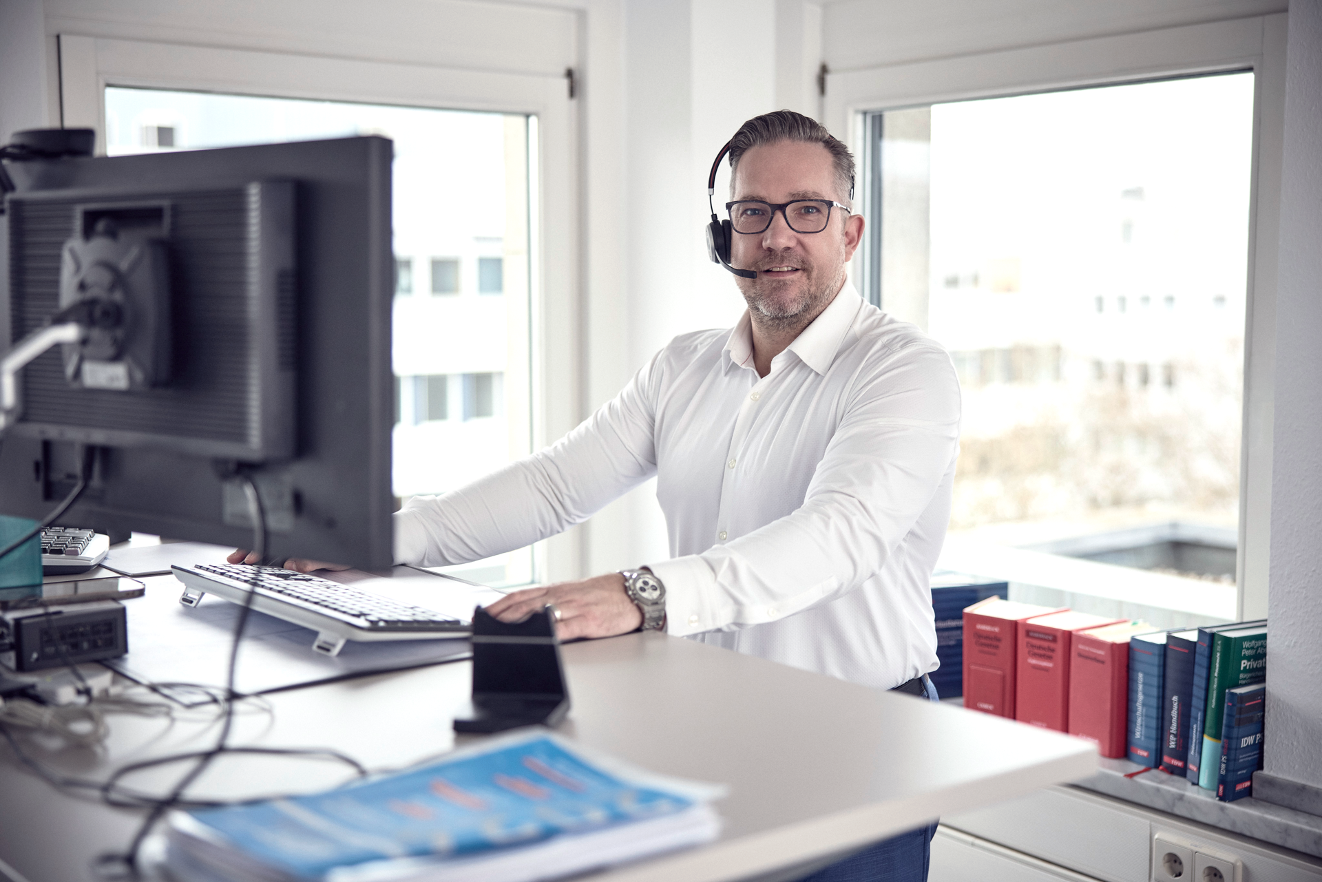 Ein Mann in einem weißen Hemd sitzt an einem Schreibtisch in einem modernen Büro, trägt ein Headset und lächelt in die Kamera. Im Hintergrund sind Bücherregale mit verschiedenen Gesetzestexten zu sehen und ein Computerbildschirm befindet sich direkt vor ihm.