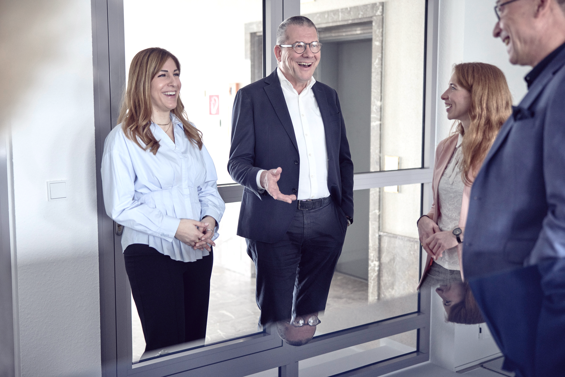 Eine Gruppe von vier Personen steht in einem modernen Büro, alle lächeln und scheinen sich amüsant zu unterhalten. Eine Frau trägt ein hellblaues Oberteil, der Mann in der Mitte hat eine Brille und spricht, während zwei weitere Frauen, eine in einem blassen Rosa und eine mit langen, roten Haaren, aufmerksam zuhören.