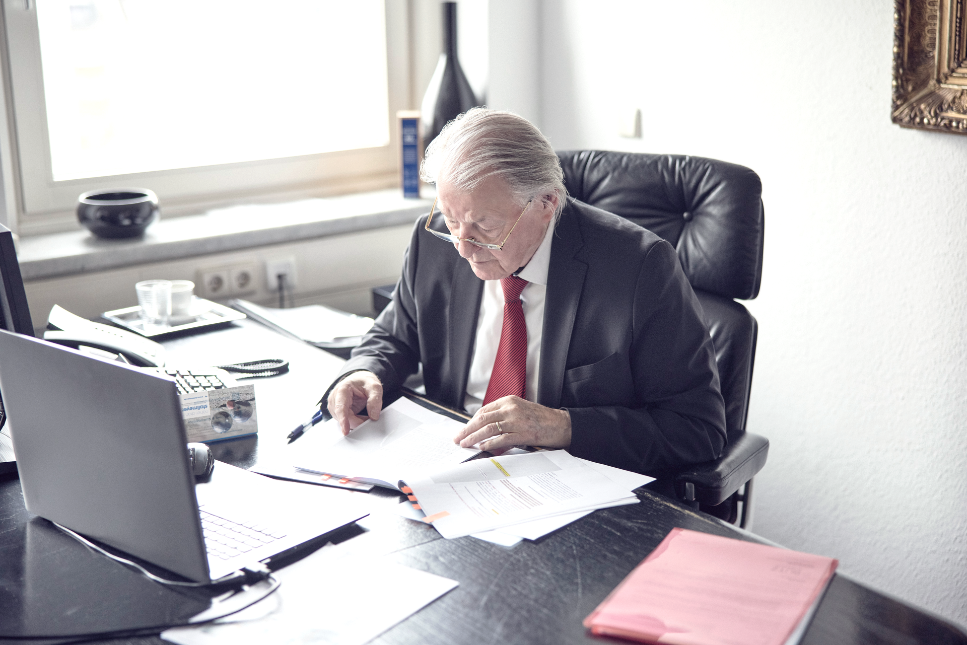 Ein älterer Mann mit grauem Haar und Brille sitzt an einem Schreibtisch und studiert Dokumente. Er trägt einen Anzug mit roter Krawatte und ist von Büromaterialien umgeben, während er auf einen Laptop schaut. Hinter ihm befindet sich ein helles Fenster.