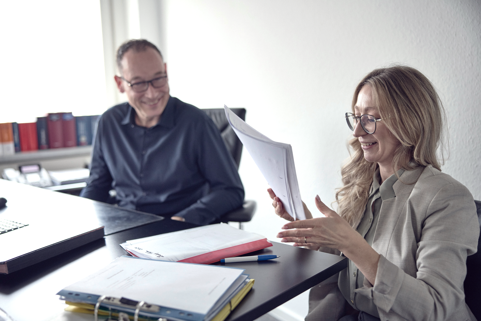 Eine lächelnde Frau mit Brille zeigt ein Dokument während eines Gesprächs mit einem Mann in einem modernen Büroraum, umgeben von Aktenordnern und einem Laptop.