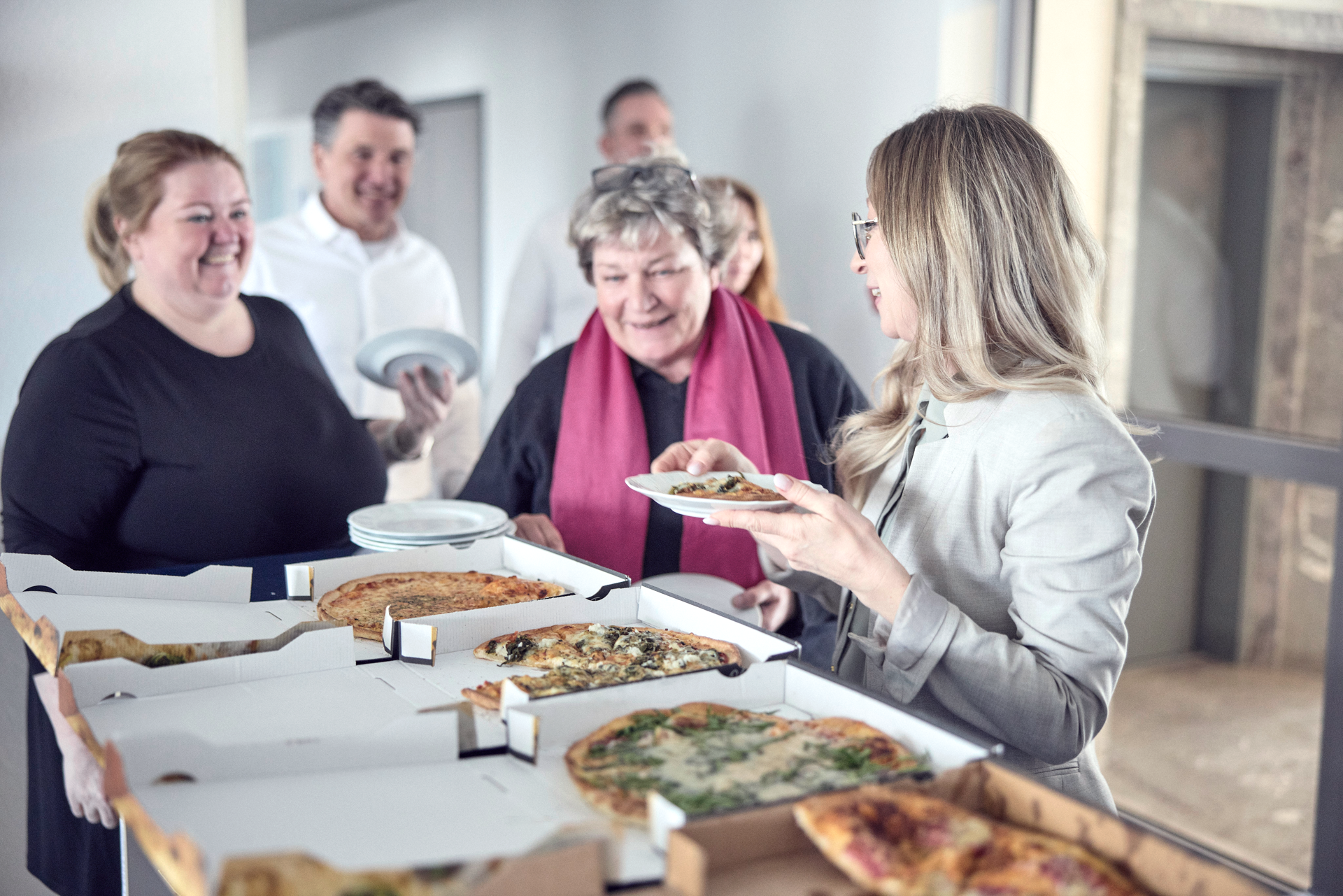Eine Gruppe von Personen steht in einem Raum, während eine Frau mit blonden Haaren auf einen Teller mit Essen schaut, den sie in der Hand hält. Auf einem Tisch liegen mehrere Pizzakartons mit verschiedenen Pizzas. Im Hintergrund sind weitere Personen, die offensichtlich an der Essensausgabe teilnehmen. Die Atmosphäre wirkt freundlich und gesellig.