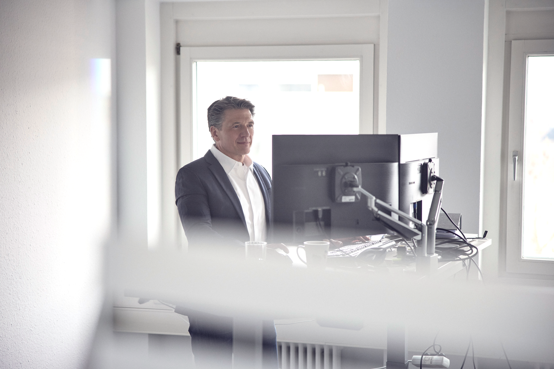 Ein Mann in einem dunklen Anzug steht an einem Schreibtisch mit einem Computerbildschirm, während er durch ein Fenster schaut. Im Hintergrund sind unscharfe Fenster und eine helle Umgebung zu sehen.