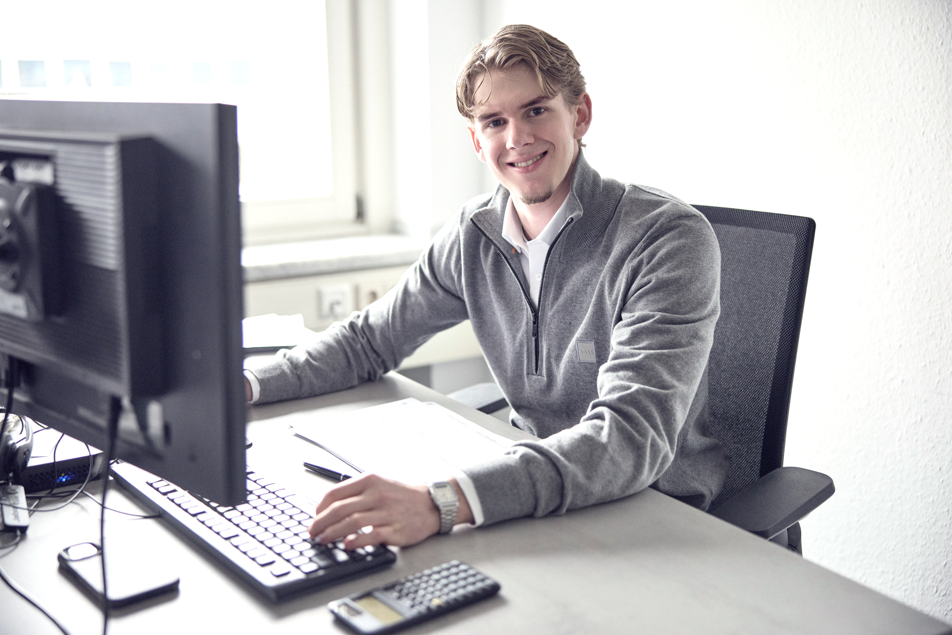 Ein junger Mann sitzt an einem Schreibtisch, lächelt in die Kamera und arbeitet an einem Computer. Auf dem Tisch liegen ein Notizblock und ein Taschenrechner.