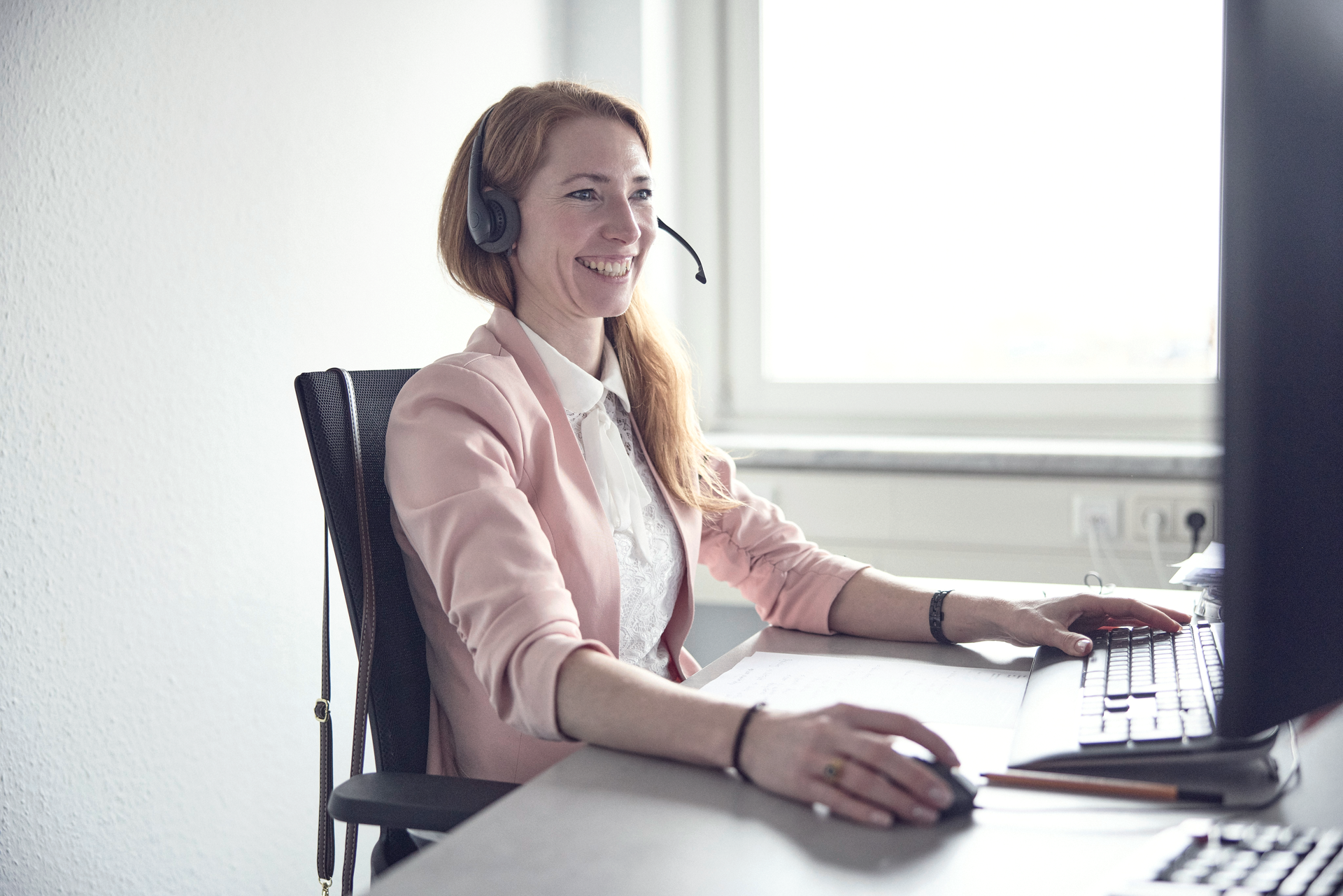 Eine lächelnde Frau mit Kopfhörern sitzt an einem Schreibtisch und arbeitet am Computer, während sie in einem hellen, freundlichen Raum sitzt.