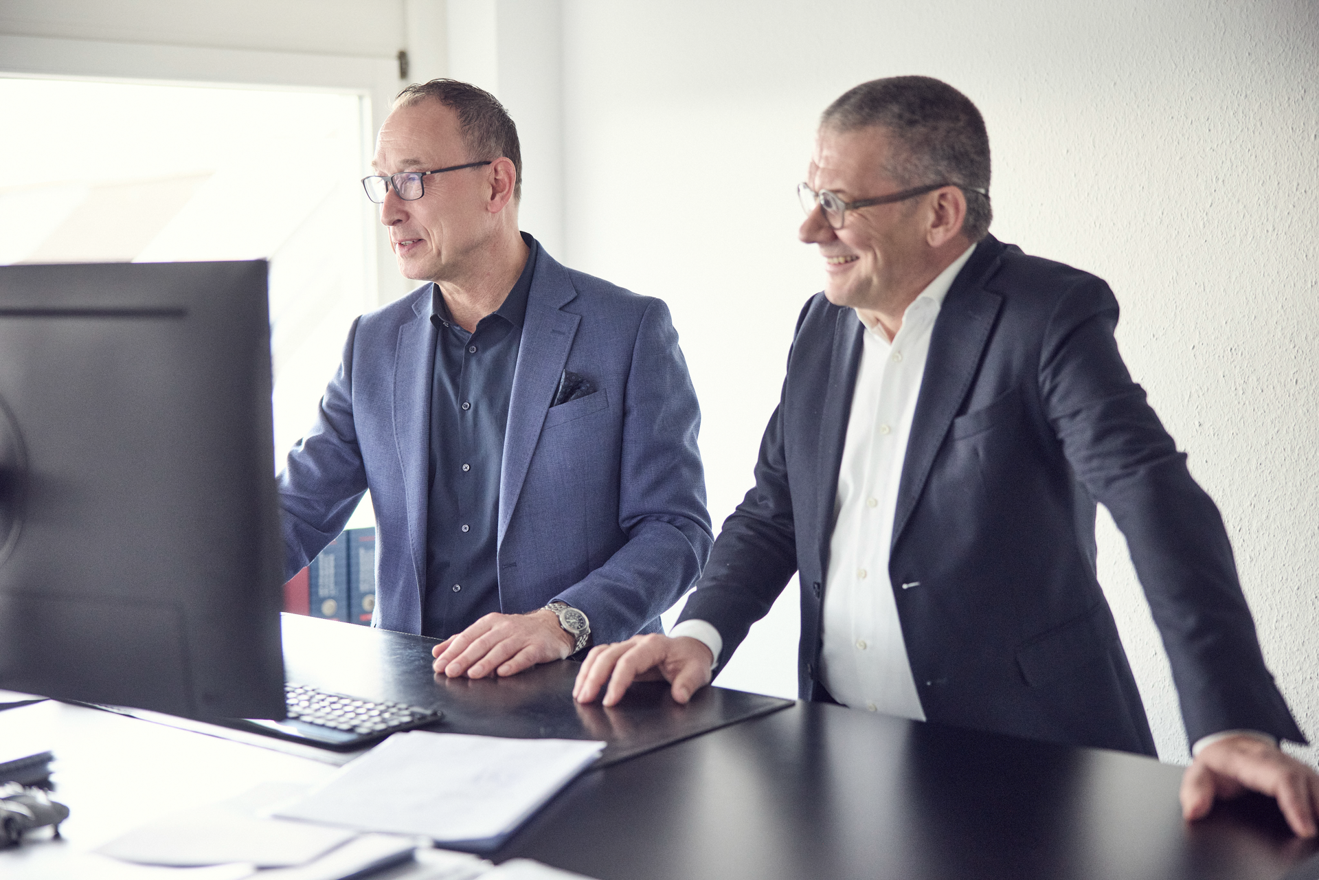 Zwei Männer in Anzügen stehen an einem Schreibtisch und schauen auf einen Computerbildschirm, während sie miteinander sprechen und lächeln.