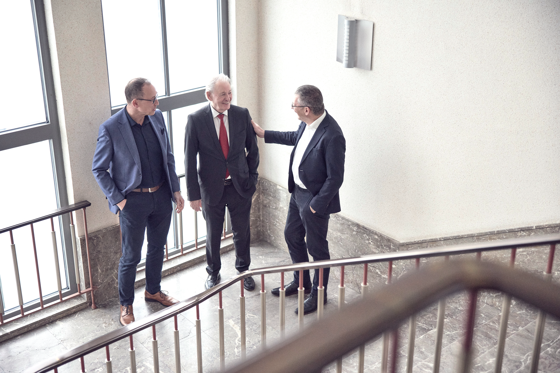 Drei Männer stehen auf einer Treppe und unterhalten sich freundlich, während sie an einem Geländer lehnen. Die Umgebung ist gut beleuchtet mit großen Fenstern, die Tageslicht hereinlassen.
