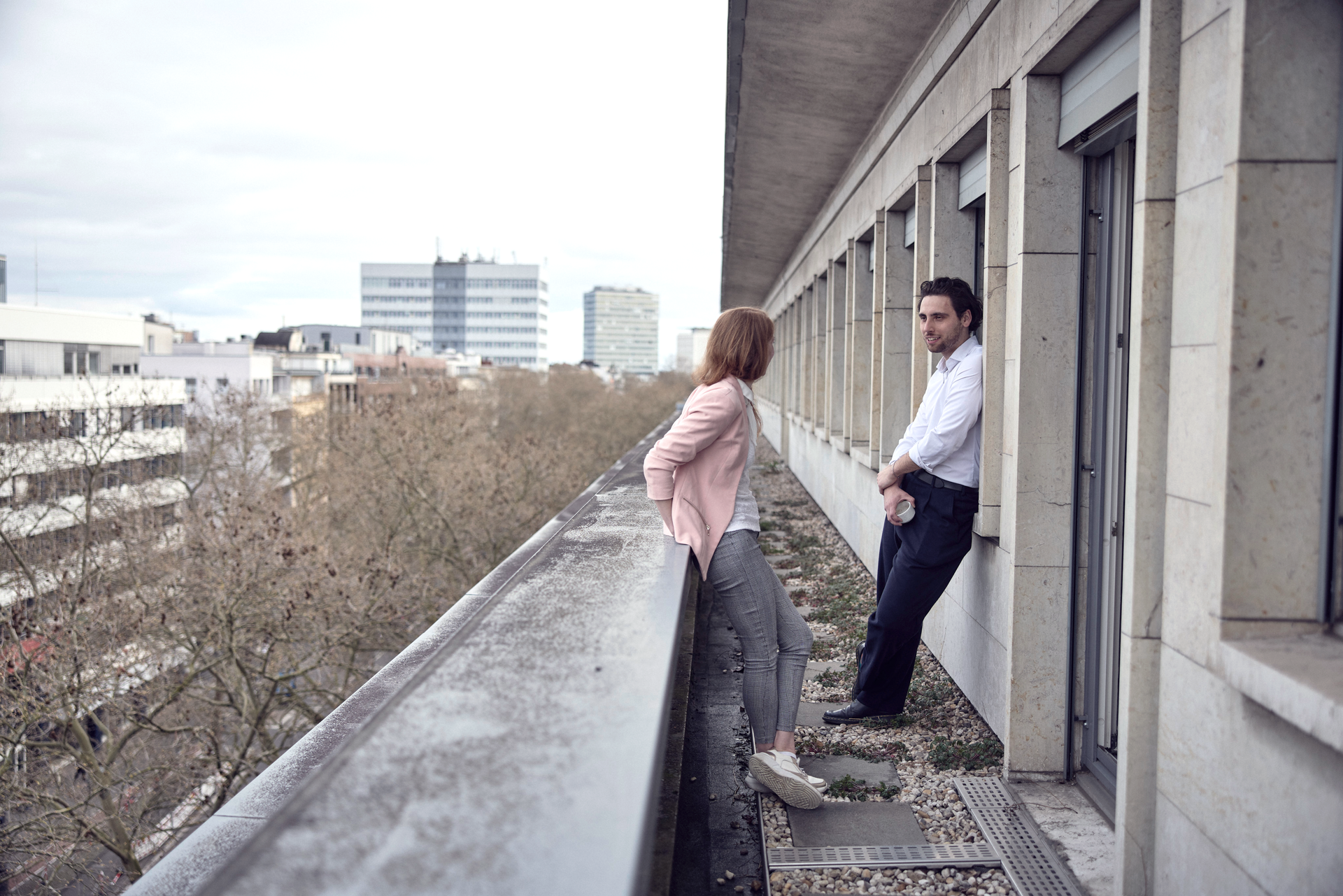 Ein Mann im weißen Hemd lehnt an einem Geländer und schaut zu einer Frau in einem rosa Blazer, die neben ihm steht und ebenfalls nach draußen blickt, während sie sich auf einem Balkon in einer städtischen Umgebung befinden.